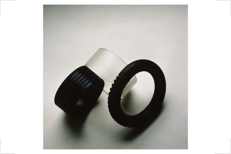 Gestaltungslinie Plastschmuck, Design Jürgen Raudis, 1986/87, Modelle, nicht realisiert, Auftraggeber: Plastverarbeitende Industrie (AiF / Handelsunternehmen Exquisit), Fotos/Zeichnungen: Jürgen Raudis. Armreifen, links Plast massiv, umlaufend 0,25mm vertiefte Profilstreifen mattiert, in Gegenrichtung erhaben laufendes Profil glänzend, Armreifen rechts, Einzelglieder mittels Steckprinzip verbunden, kombiniert mit halbseitig geschlossener Form oder Einzelelemente durchgehend zu Armreifen bzw. Halsschmuck gereiht, Variabilität durch Material, Farb- und Oberflächengestaltung.